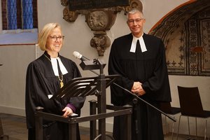 Zwei Menschen in geistlicher Kleidung stehen nebeneinander, die Frau spricht in ein Mikrofon