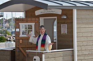 Eine Frau strahlt einladend vor der geöffneten Tür der Schäferwagenkirche in die Kamera